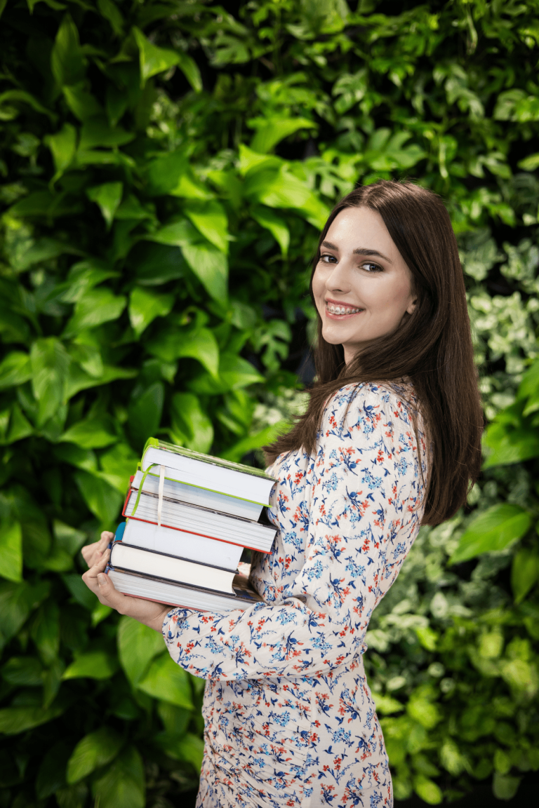 Who we are | WSB Merito Gdańsk University: Zdjęcie młodej studentki z aparatem na zębach. Ma brązowe włosy do ramion i delikatną białą sukienkę w małe kwiatki. Trzyma w ręku stertę książek. Pozuje na tle zielonej ściany z kwiatami