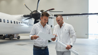 Obsługa ruchu lotniczego | Studia I stopnia w Gdańsku | Uniwersytet WSB Merito w Gdańsku: Dwóch ubranych na jasno mężczyzn patrzy na kartkę i znajdują się w przestrzeni, gdzie jest samolot
