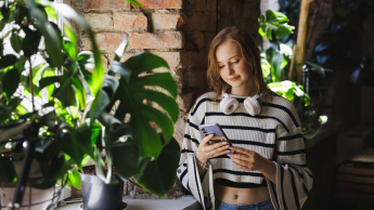 Digital Marketing | Studia II stopnia ze studiami podyplomowymi  | Uniwersytet WSB Merito w Gdańsku: Studentka z komórką stoi przy oknie przy zielonym parapecie