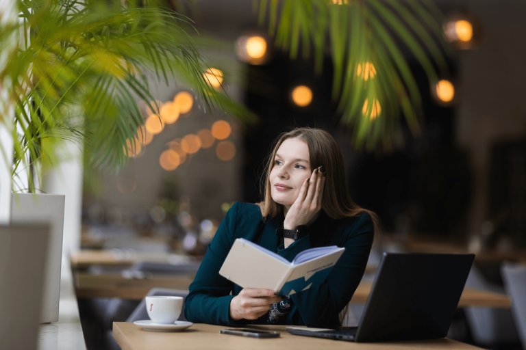 Biblioteka | Uniwersytet WSB Merito w Gdańsku: Portret młodej, naturalnej kobiety o delikatnych rysach i włosach. Siedzi przy biurku w kawiarni oparta dłonią o głowę, w drugiej ręce trzyma zeszyt z logotypem UWSB Merito