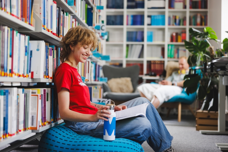 Specjalista kadr i płac z elementami technik coachingu | Studia II stopnia w Gdańsku | Uniwersytet WSB Merito w Gdańsku: Studentka w bibliotece z książką na kolanach