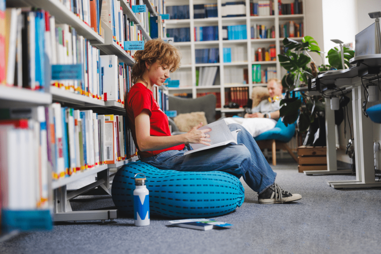 Marketing i sprzedaż | Studia II stopnia ze studiami podyplomowymi w Gdańsku | Uniwersytet WSB Merito w Gdańsku: Studentka w bibliotece siedzi na puffie i przegląda książkę 