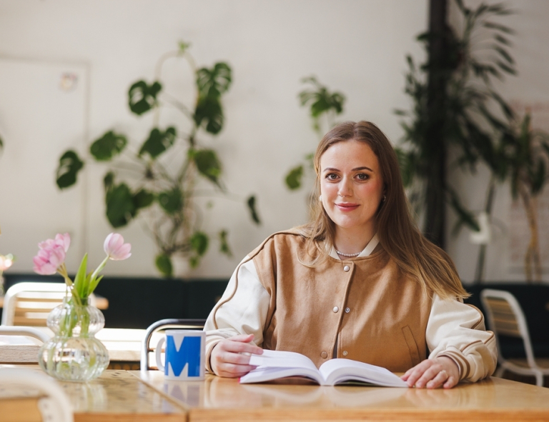 Komunikacja cyfrowa i media kreatywne | Studia II stopnia ze studiami podyplomowymi w Gdańsku | Uniwersytet WSB Merito w Gdańsku: Studentka w kawiarni z książką i kubkiem z literą M