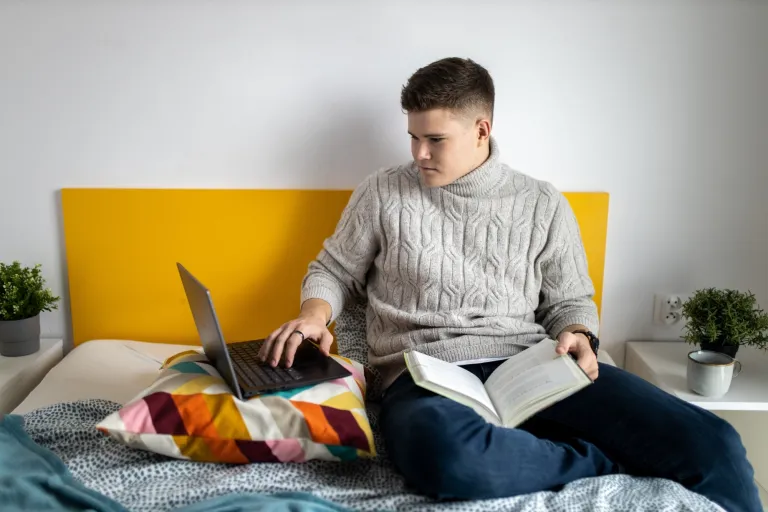 Zarządzanie projektami i usługami IT | Studia II stopnia w Gdańsku | Uniwersytet WSB Merito w Gdańsku: Student siedzi w łóżku z laptopem i ma na kolanach książkę