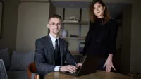 Programista Python Developer | Studia podyplomowe w Gdyni | Uniwersytet WSB Merito Gdynia: Zdjęcie osób w średnim wieku. Mężczyzna w garniturze siedzi przy laptopie, obok niego stoi kobieta ubrana na czarno