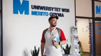 Kadry i płace | Studia I stopnia w Gdyni | Uniwersytet WSB Merito Gdynia: Student na korytarzu przy logo Uczelni