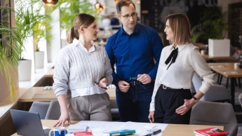 Sztuczna inteligencja w digital marketingu | Studia podyplomowe w Gdyni | Uniwersytet WSB Merito Gdynia: Rozmowa trzech osób stojących przy biurku, omawiają jakiś projekt