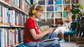 Psychologia reklamy i komunikacji | Studia podyplomowe w Gdyni | Uniwersytet WSB Merito Gdynia: Studentka ze słuchawkami w uszach, książką na kolanach i telefonem w ręku