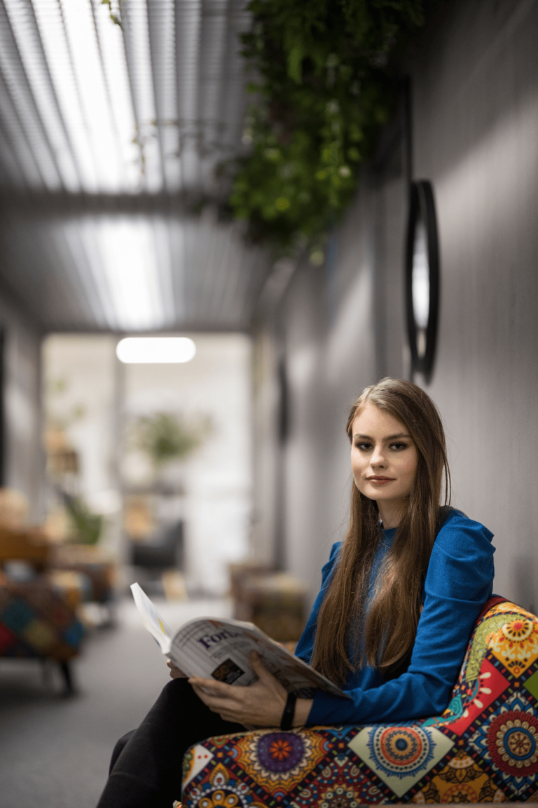 Transport autonomiczny i bezzałogowy | Studia I stopnia w Gdyni | Uniwersytet WSB Merito Gdynia: W szarej przestrzeni w fotelu siedzi młoda studentka z gazetą na kolanach. Siedzi w patchworkowym kolorowym fotelu