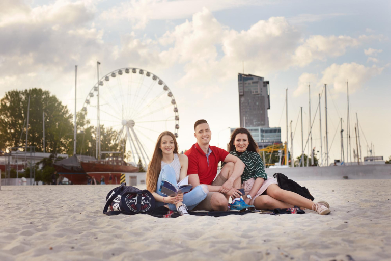 Tłumaczenia – język angielski | Studia I stopnia w Gdyni | Uniwersytet WSB Merito Gdynia: Fotografia robiona na plaży w Gdyni, z tyłu widać wieżowiec i koło widokowe. Na plaży siedzą dwie studentki i student