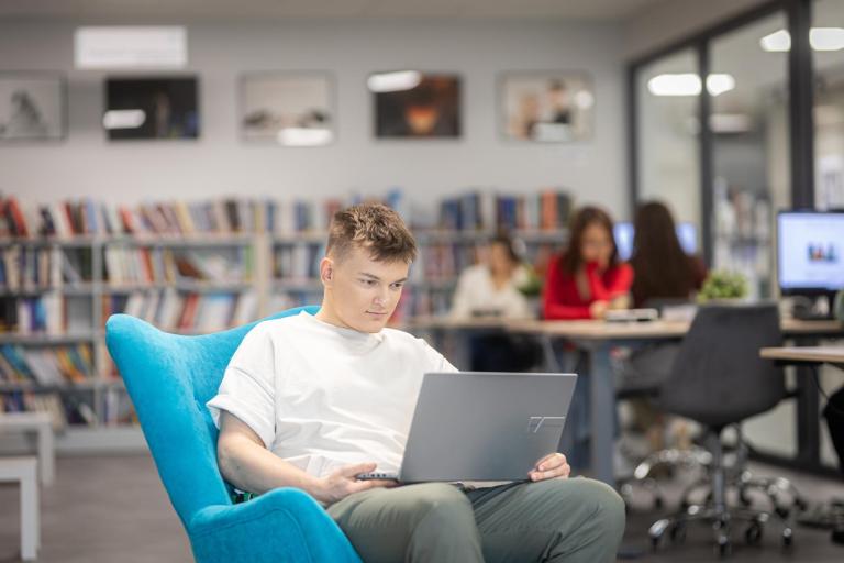 Negocjacje i mediacje | Studia I stopnia w Gdyni | Uniwersytet WSB Merito Gdynia: Student w przestrzeni biblioteki siedzi na fotelu i na kolanach trzyma laptopa