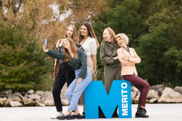 Psychologia w biznesie | Studia I stopnia w Gdyni | Uniwersytet WSB Merito Gdynia: Zdjęcie grupowe studentek, które robią sobie selfie na zewnątrz. Opierają się one o sygnet litery "m"