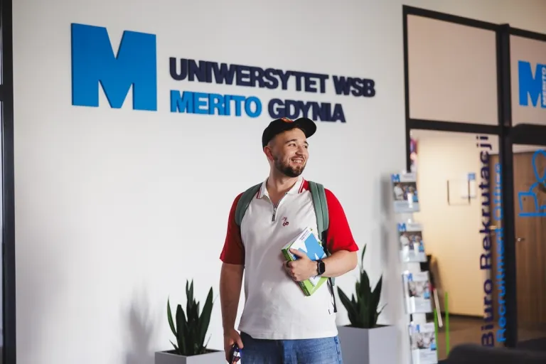 Zarządzanie | Studia I stopnia w Gdyni | Uniwersytet WSB Merito Gdynia: Student na korytarzu przy logo Uczelni