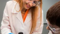 Zarządzanie i kontrola w podmiotach leczniczych | Studia podyplomowe w Łodzi | Uniwersytet WSB Merito Łódź: Kobieta w laboratorium w białym fartuchu i okularach ochronnych pokazuje dziecku, jak korzystać z miernika pH, wykonując doświadczenie przy stole z próbkami.