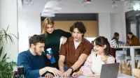 Zarządzanie jakością | Studia podyplomowe w Łodzi | Uniwersytet WSB Merito Łódź: Czworo młodych ludzi współpracuje przy stole, analizując kolorowe materiały i korzystając z laptopa, w nowoczesnym wnętrzu sprzyjającym nauce i kreatywnej pracy zespołowej.