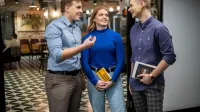 Zarządzanie zespołami rozproszonymi | Uniwersytet WSB Merito Łódź: Trójka studentów rozmawia ze sobą w przestrzeni coworkingowej, trzymając książki i uśmiechając się podczas dyskusji.