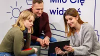 Troje młodych ludzi wspólnie ogląda coś na tablecie, siedząc w nowoczesnym wnętrzu z napisem „Miło Cię widzieć” w tle.