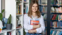 Psychologia sądowo - penitencjarna | Studia jednolite magisterskie w Łodzi | Uniwersytet WSB Merito Łódź: Młoda kobieta w okularach, trzymająca podręcznik w bibliotece, uśmiecha się stojąc przy regale z książkami.