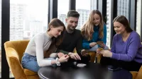 Bezpieczeństwo i higiena pracy | Studia podyplomowe w Łodzi | Uniwersytet WSB Merito Łódź: Cztery młode osoby siedzą przy okrągłym stole w jasnym pomieszczeniu z dużymi oknami, skupione na wspólnej grze karcianej i uśmiechające się.