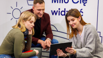 Troje młodych ludzi wspólnie ogląda coś na tablecie, siedząc w nowoczesnym wnętrzu z napisem „Miło Cię widzieć” w tle.
