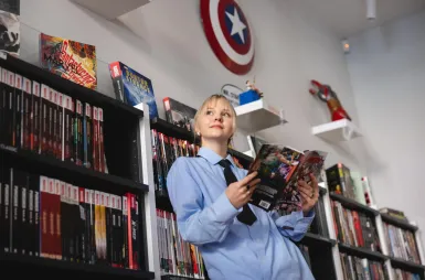 Organizacje i koła naukowe | Uniwersytet WSB Merito Łódź: Kobieta w niebieskiej koszuli czyta komiks, otoczona półkami pełnymi komiksów i dekoracjami Marvela.