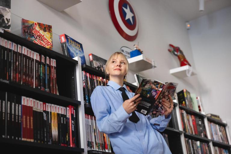 Stypendia i zniżki | Uniwersytet WSB Merito Łódź: Kobieta w niebieskiej koszuli czyta komiks, otoczona półkami pełnymi komiksów i dekoracjami Marvela.