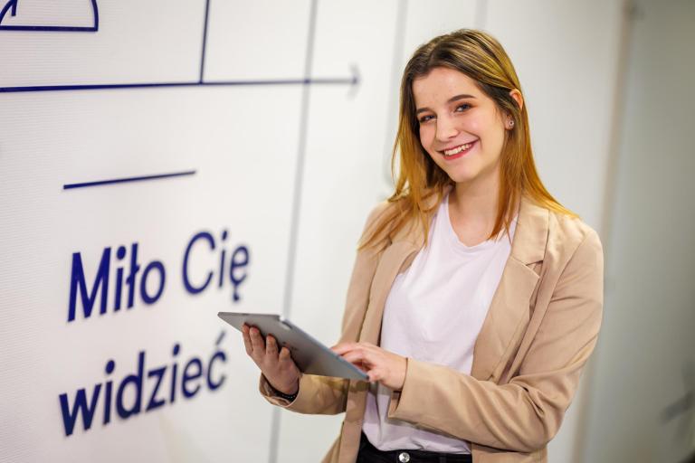 Nowe media | Studia I stopnia w Łodzi | Uniwersytet WSB Merito Łódź: Studentka z tabletem na tle napisu "Miło Cię widzieć"