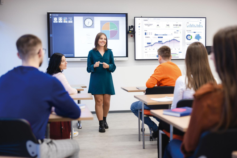 Zarządzanie projektami | Studia II stopnia w Łodzi | Uniwersytet WSB Merito Łódź: Młoda kobieta prowadzi prezentację w nowoczesnej sali lekcyjnej, podczas gdy studenci uważnie ją słuchają, śledząc dane na ekranach multimedialnych.