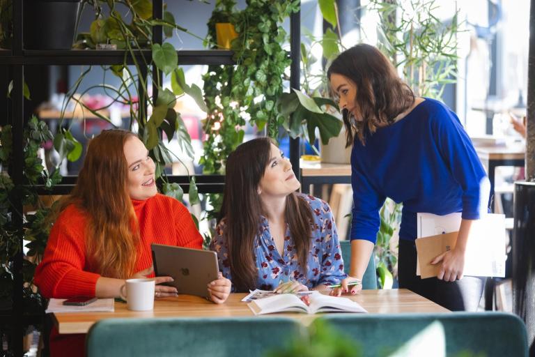 Zarządzanie przedsiębiorstwem | Studia I stopnia w Łodzi | Uniwersytet WSB Merito Łódź: Trzy kobiety pracują wspólnie przy stole w zielonej, nowoczesnej przestrzeni — rozmowa, notatki i tablet.