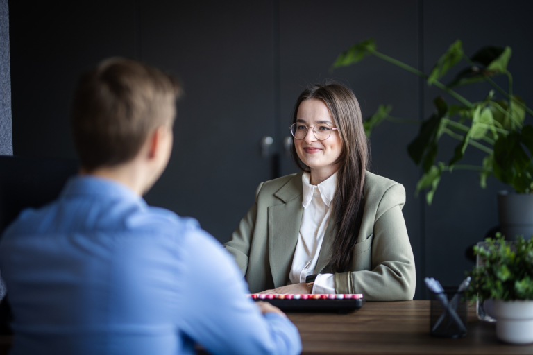 Menedżer zarządzania zakupami | Studia podyplomowe w Łodzi | Uniwersytet WSB Merito Łódź: Kobieta w okularach i eleganckim stroju uśmiecha się podczas rozmowy z mężczyzną w jasnoniebieskiej koszuli przy biurku w nowoczesnym biurze.
