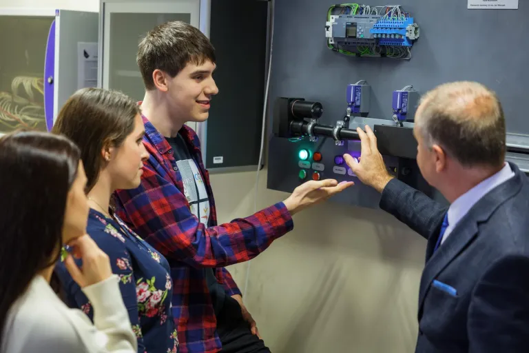 Grupa młodych osób słucha mężczyzny w garniturze, który tłumaczy działanie panelu sterującego z przyciskami i elektroniką w laboratorium technicznym.
