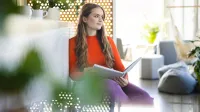 Psychologia biznesu i rozwoju osobistego | Studia jednolite magisterskie w Opolu | Uniwersytet WSB Merito Opole: Główna grafika: kobieta siedząca w kawiarni, spoglądająca w bok, trzymająca w dłoni książkę. W tle wystrój kawiarni. 