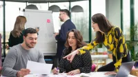 Psychologia reklamy i komunikacji | Studia podyplomowe w Opolu | Uniwersytet WSB Merito Opole: Główna grafika: Opole - studia podyplomowe - Psychologia reklamy i komunikacji - Grupa osób pracująca nad projektem, w tle dwie osoby piszące na białej tablicy