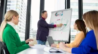 Programista JavaScript z wykorzystaniem AI | Studia podyplomowe w Opolu | Uniwersytet WSB Merito Opole: Główna grafika: Opole - studia podyplomowe - Programista JavaScript z wykorzystaniem AI - Grupa osób słuchająca mężczyzny prezentującego dane przy białej tablicy