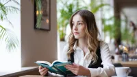 Księgowość i podatki w praktyce | Studia podyplomowe w Opolu | Uniwersytet WSB Merito Opole: Główna grafika: Opole - studia podyplomowe - Księgowość i podatki w praktyce - Kobieta siedząca w kawiarni, trzyma książkę
