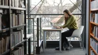 Księgowość i podatki w praktyce | Studia podyplomowe w Opolu | Uniwersytet WSB Merito Opole: Główna grafika: Opole - studia podyplomowe - Księgowość i podatki w praktyce - Kobieta korzystająca z laptopa, siedzi przy stoliku w bibliotece