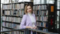Mediacje sądowe i pozasądowe     | Studia podyplomowe w Opolu | Uniwersytet WSB Merito Opole: główna grafika: Opole - studia podyplomowe - mediacje sądowe i pozasadowe - kobieta w jasnej marynarce stoi przy barierce a za sobą ma regały z książkami
