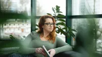 Terapia uzależnień | Studia podyplomowe w Opolu | Uniwersytet WSB Merito Opole: Główna grafika: Opole - studia podyplomowe - Terapia uzależnień - uczestniczka studiów podyplomowych siedzi na kanapie z książką w rękach i spogląda za okno