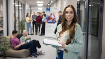 Psychodietetyka | Studia I stopnia w Opolu | Uniwersytet WSB Merito Opole: Główna grafika: Opole - psychodietetyka - uśmiechnięta studentka stojąca na korytarzu, trzyma książkę