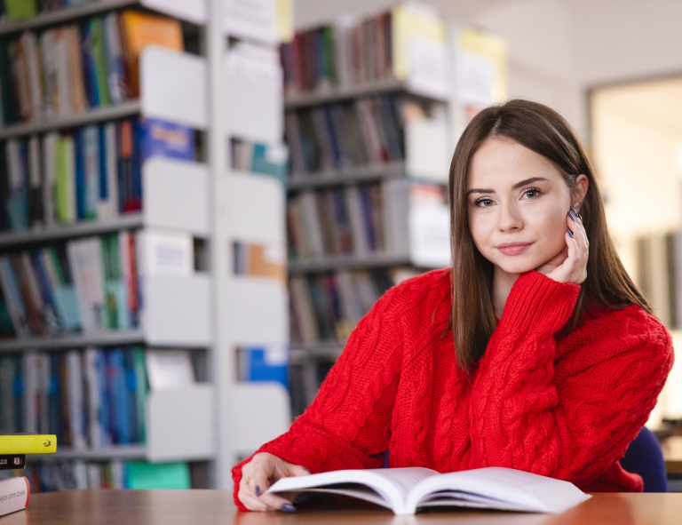 Pedagogika resocjalizacyjna z socjoterapią i kryminologią | Studia II stopnia w Opolu | Uniwersytet WSB Merito Opole: Dziewczyna w bibliotece, ma przed sobą otwartą książkę, jest ubrana w czerwony sweter 