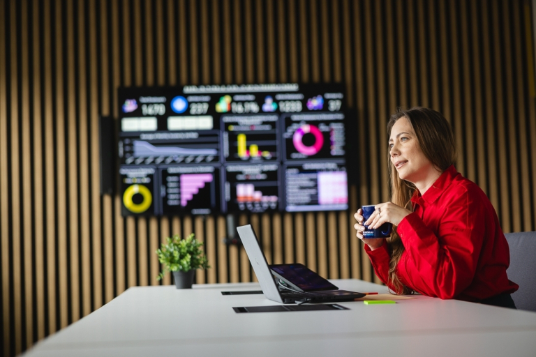Artificial intelligence. Machine learning | Studia podyplomowe w Opolu | Uniwersytet WSB Merito Opole: Kobieta korzystająca z laptopa, trzyma kubek kawy. Za nią duży ekran, na którym wyświetlają się dane i wykresy