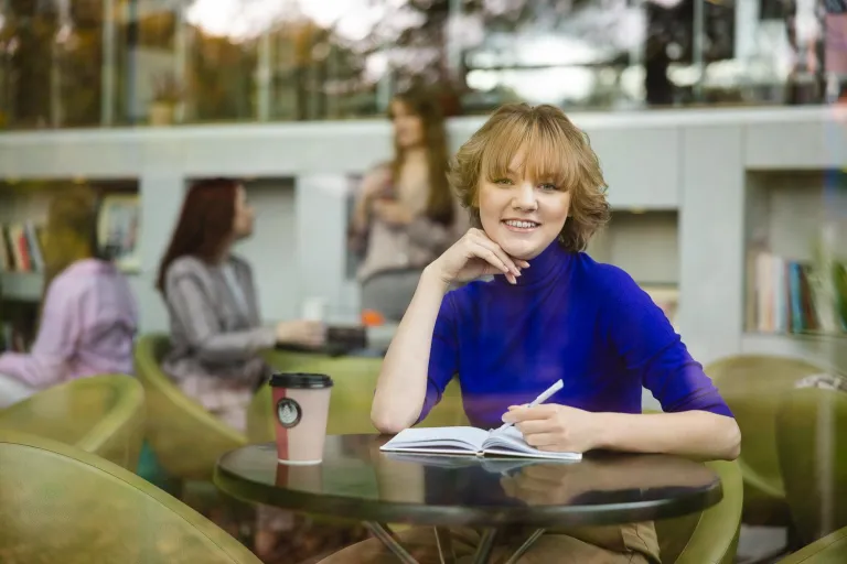Administracja i bezpieczeństwo wewnętrzne | Studia I stopnia w Opolu | Uniwersytet WSB Merito Opole: Uśmiechnięta studentka. Siedzi w kawiarni. Pisze w notatniku