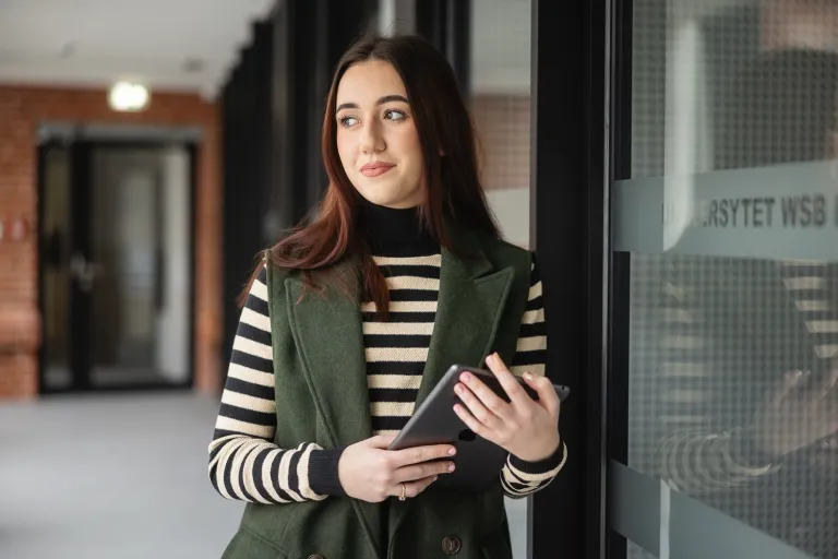 Dietetyka sportowa | Studia II stopnia ze studiami podyplomowymi  | Uniwersytet WSB Merito Opole: Uczestniczka studiów podyplomowych stoi i opera się plecami o półkę w bibliotece, trzymając w ręce książki