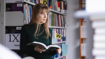 Rachunkowość od podstaw | Studia podyplomowe w Szczecinie | Uniwersytet WSB Merito Szczecin: Studentka Uniwersytetu WSB Merito spogląda za okno, przeglądając książkę w bibliotece.