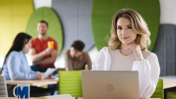 Organizacja i zarządzanie oświatą | Studia podyplomowe w Szczecinie | Uniwersytet WSB Merito Szczecin: Studentka pracuje przy laptopie w nowoczesnej przestrzeni z innymi osobami w tle.