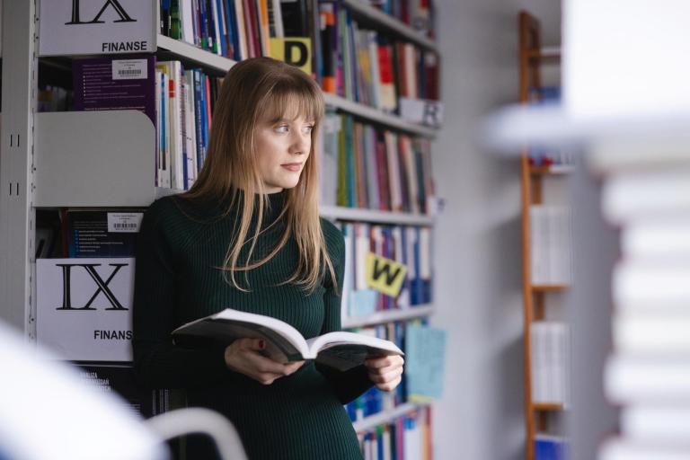 Wpływ społeczny | Uniwersytet WSB Merito Szczecin: Studentka Uniwersytetu WSB Merito spogląda za okno, przeglądając książkę w bibliotece.
