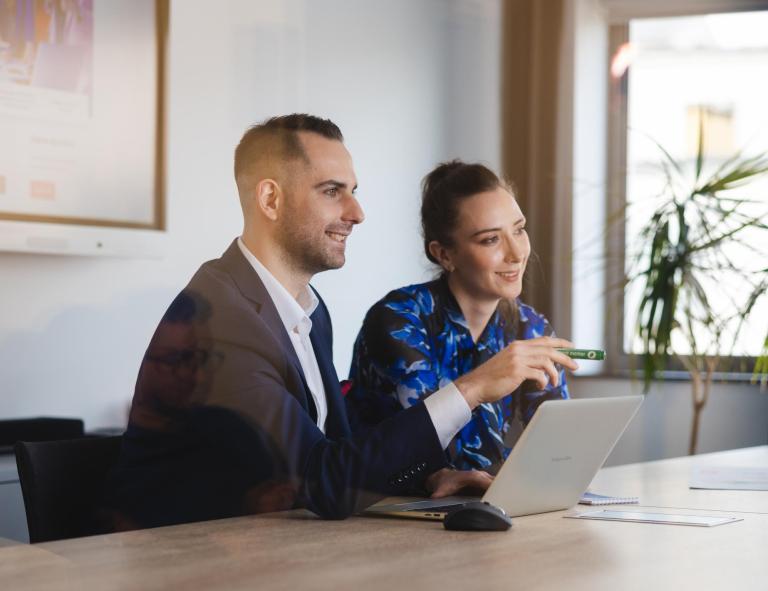 Business Intelligence – analiza danych | Studia podyplomowe w Szczecinie | Uniwersytet WSB Merito Szczecin: Dwie osoby siedzą przy stole w sali konferencyjnej, pracując wspólnie przy laptopie.
