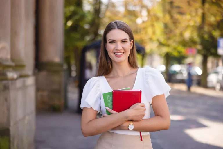 Administracja i bezpieczeństwo wewnętrzne | Studia II stopnia w Szczecinie | Uniwersytet WSB Merito Szczecin: Studentka stoi na tle miejskiej ulicy z zeszytami w rękach, uśmiechając się do obiektywu.